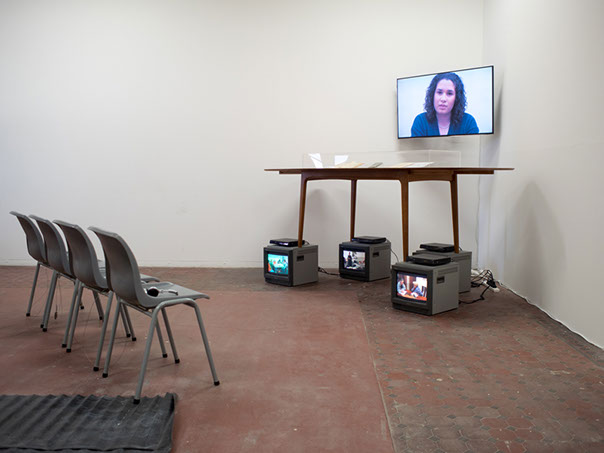 Video installation situated in a corner, 4 tv's below the table legs, a flatscreen above, identity documents sit the on the table, with 4 chairs
