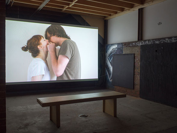 an underground dark space has a video projection of two white people who are maybe kissing ot passing air via each others mouths