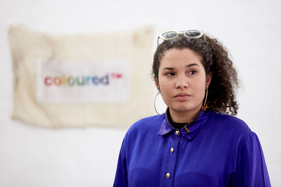 Woman with long brown curly hair tied back wearing a purple shirt with gold buttons looks to the right. In the backgroung is a tapestry artwork.