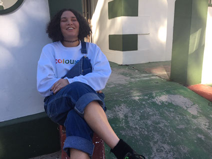 Woman wearing denim overalls sitting on a stoep laughing wearing a white jumper that reads the words in rainbow, ColouredTM