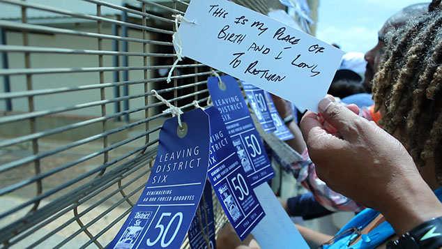 Video still of people next to tags hanging along a metal gate with a hand showing one that reads This is my Place of Birth and I long to Return.