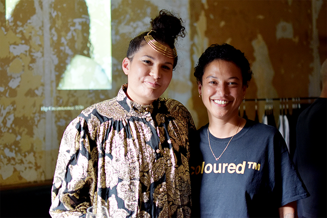 Two southern african womxn smiling at the camera, one dressed in a black and gold dress, the other a black tshirt that reads colouredtm in gold.