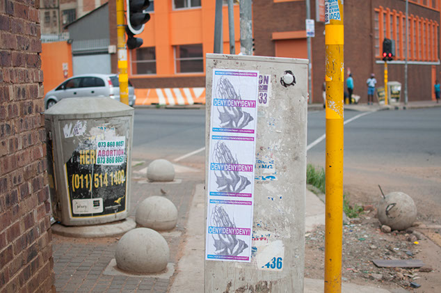 Street view of a corner in Johannesburg with paste ups on the sitde of bines and electrical boxes that look like ads, with prayer hands.