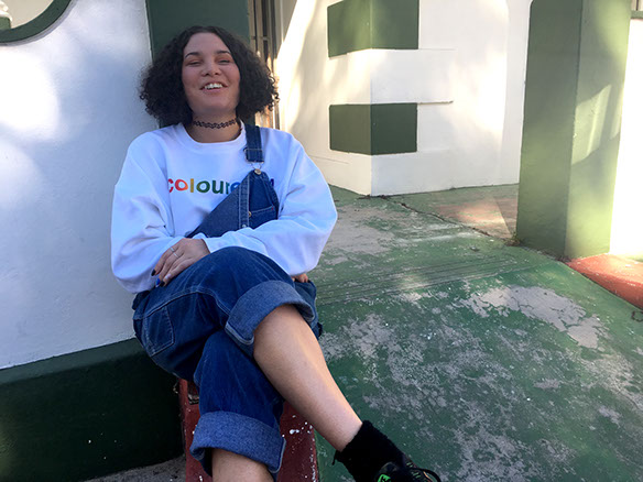 Womxn wearing a white jumper that says coloured and denim overalls sits upon a sunny stoep with laughter.