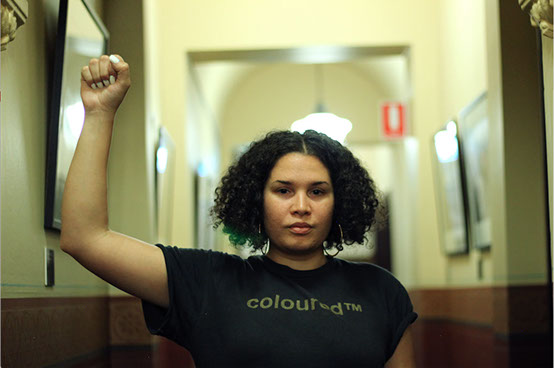Woman with curly hair has a fist raised, wearing a black tshirt reading ColouredTM in the hall of a colonial building.